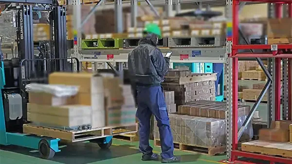 倉庫内の棚の前でピッキングを行っているところ（ハンディ端末で商品を照合し、数量を入力しているところ）